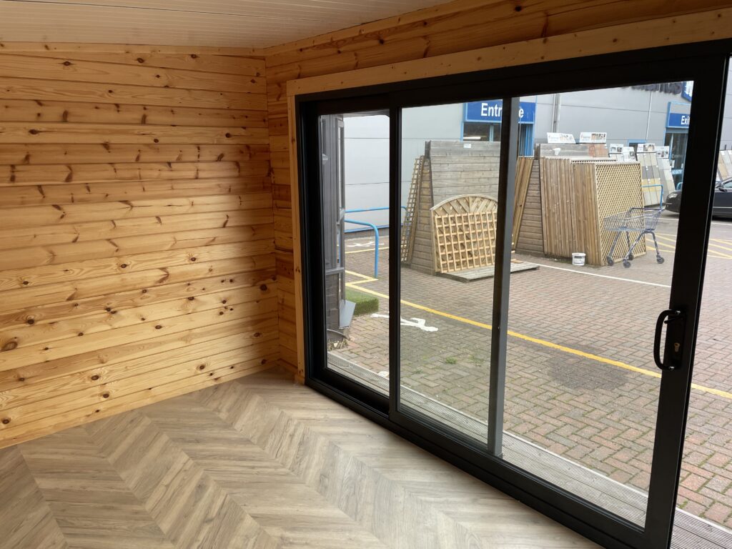 Interior of the Tiger Black Obsidian 70 mm Insulated Log Cabin at the Horsforth show site showing herringbone flooring, natural timber walls, and large sliding glass doors.