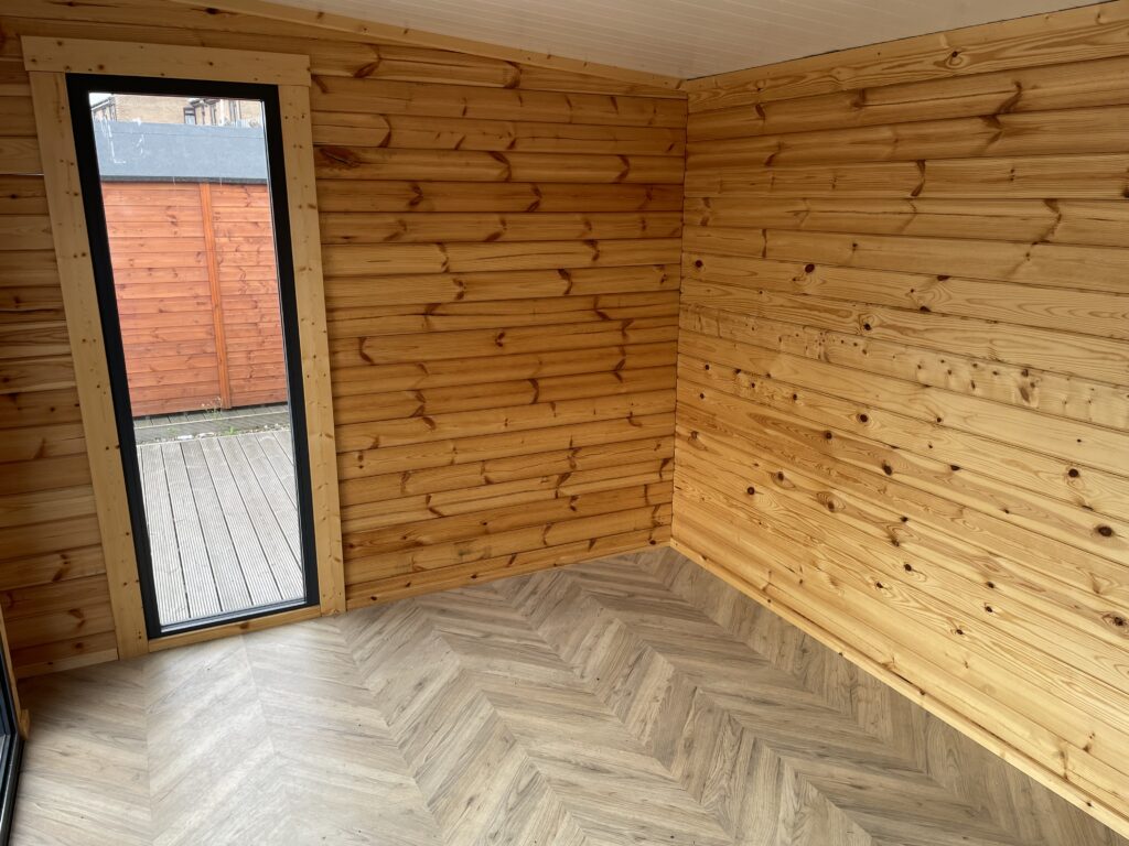 Interior corner of the Tiger Black Obsidian 70 mm Insulated Log Cabin at the Horsforth show site showing slimline window, herringbone flooring, and smooth timber wall finish.