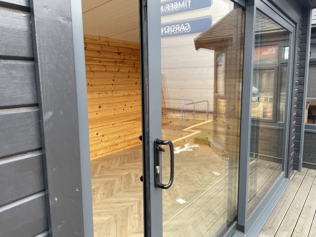 Open sliding door of the Tiger Black Obsidian 70 mm Insulated Log Cabin at the Horsforth show site showing the dark exterior, modern glazing, and warm timber interior.