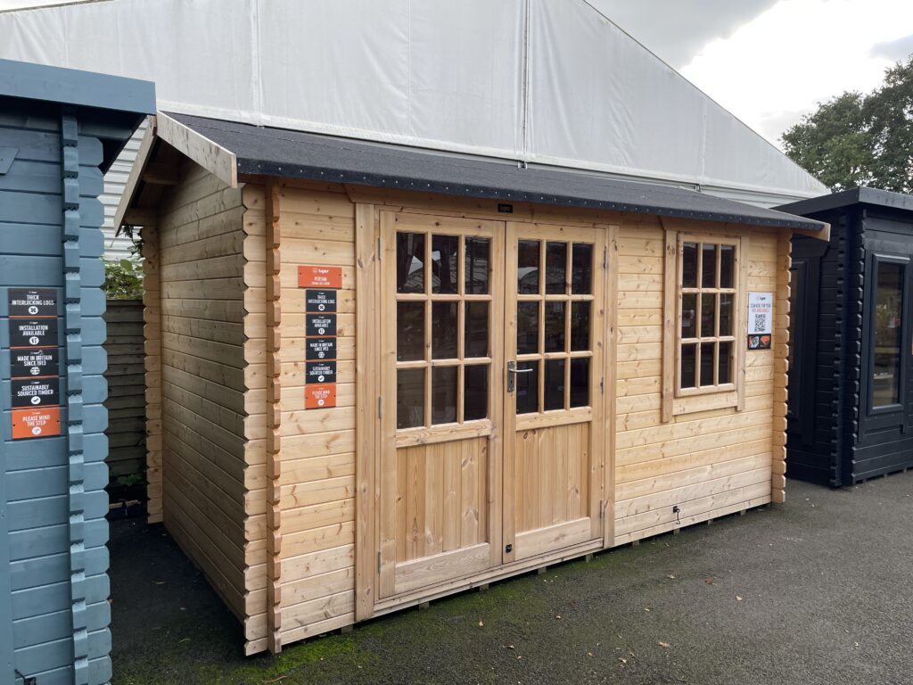 Angled front and side view of the Tiger Persian 28 mm Log Cabin at the Otley show site showing the double doors, Georgian-style window, and roof overhang.