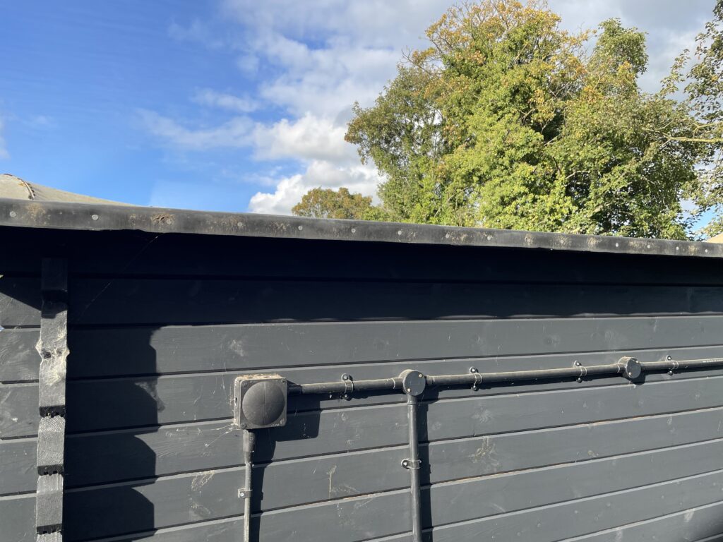 Back view of Tiger Serena 44 mm Log Cabin at Otley show site showing black cladding, flat pent roof, and outdoor electrical conduit installation.