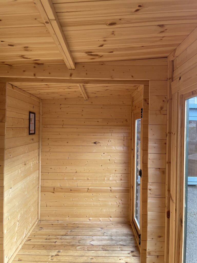 Interior view of the TigerFlex Moda 19 mm Log Cabin at the Otley show site showing the timber wall panelling, roof beams, and bright glazed doorway.